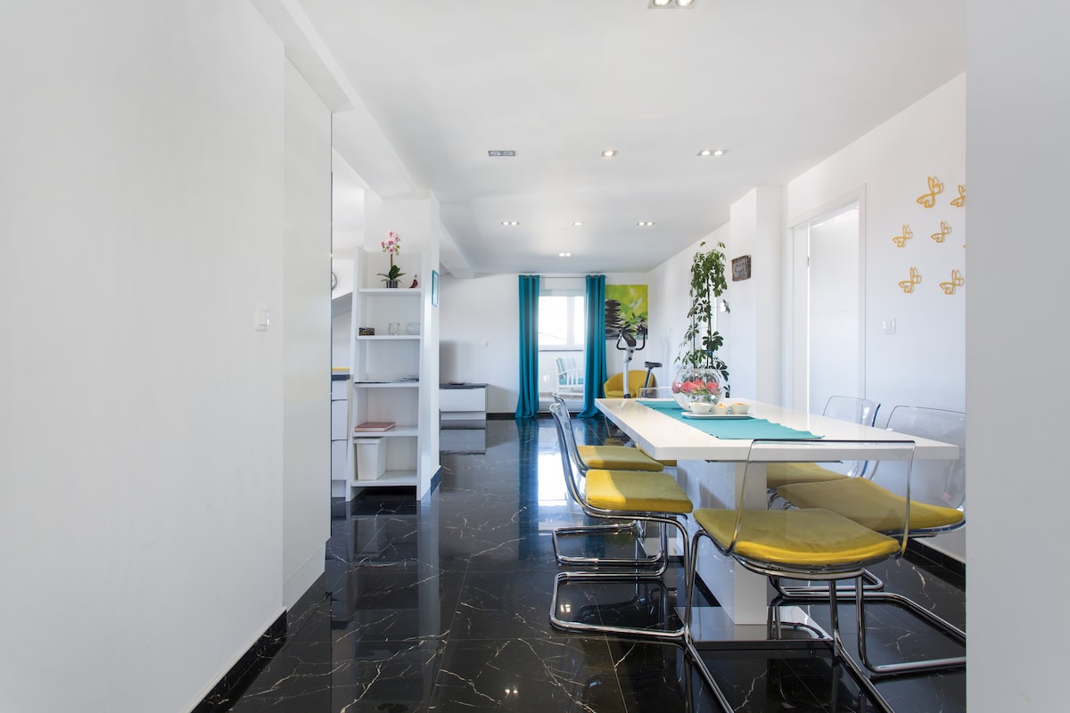 View down the apartment along a polished black marble floor with white walls, a white shelving unit on the left, the dining table with yellow-cushioned chairs on the right and turquoise curtains at the end window