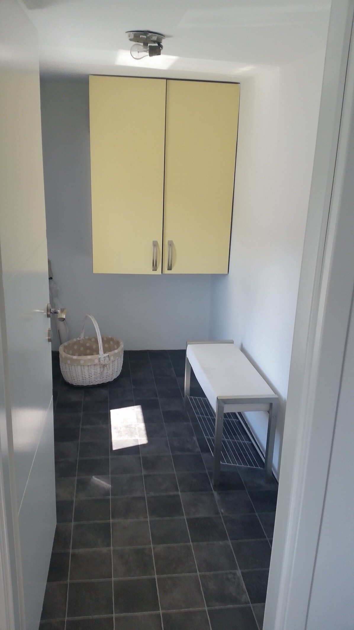 Small private entrance hall with dark grey floor tiles, a pale yellow wall cupboard, a white bench with a shoe rack beneath it and a wicker basket on the floor