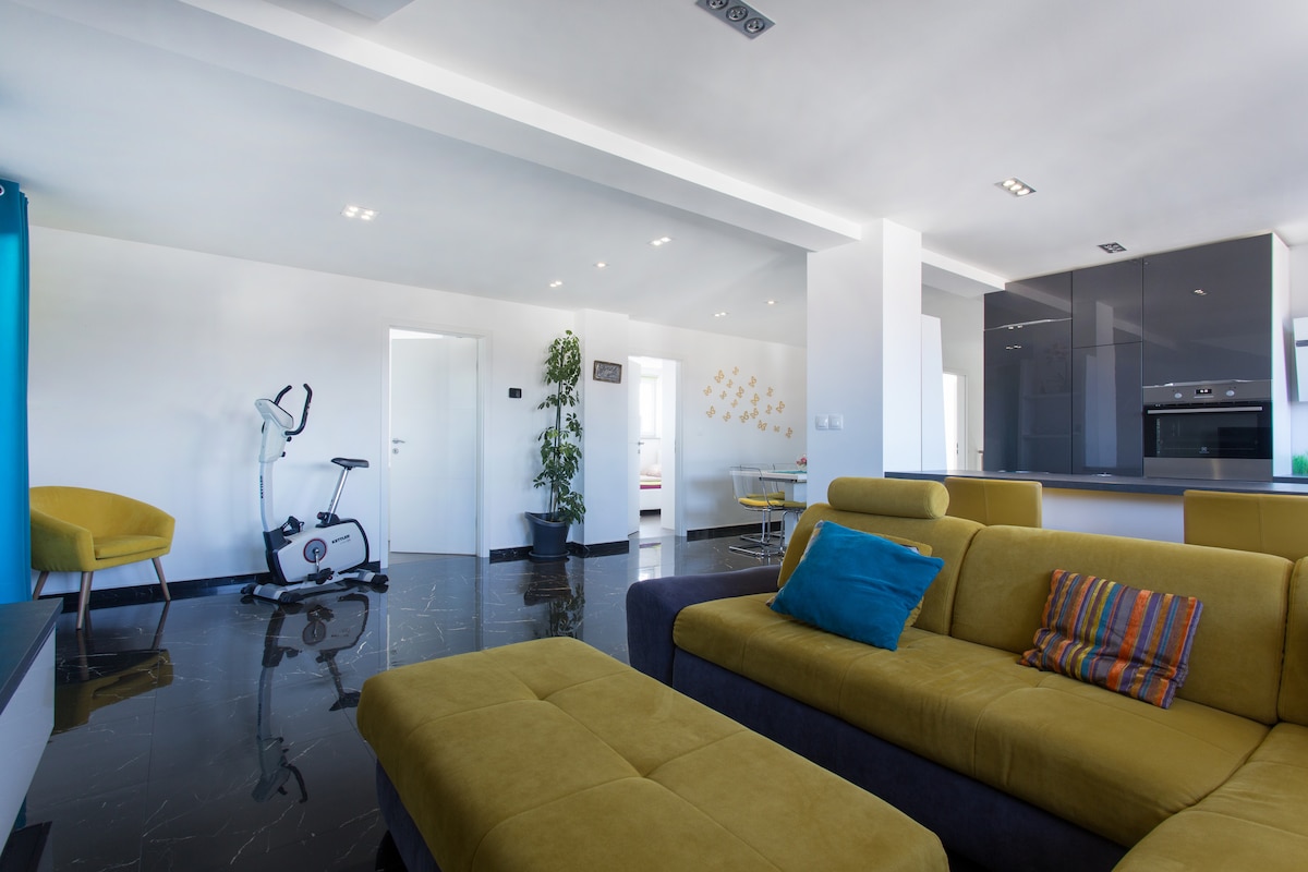 Open-plan top-floor living space with a mustard sofa and ottoman in the foreground, an exercise bike and yellow armchair against the left wall, white doors to bedrooms and the kitchen on the right
