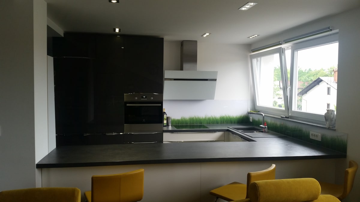 Kitchen with a black gloss column of tall units holding the oven and the fridge, a stainless chimney hood and a white angled hood, white gloss base units under a dark worktop, a corner window and yellow bar stools at the counter