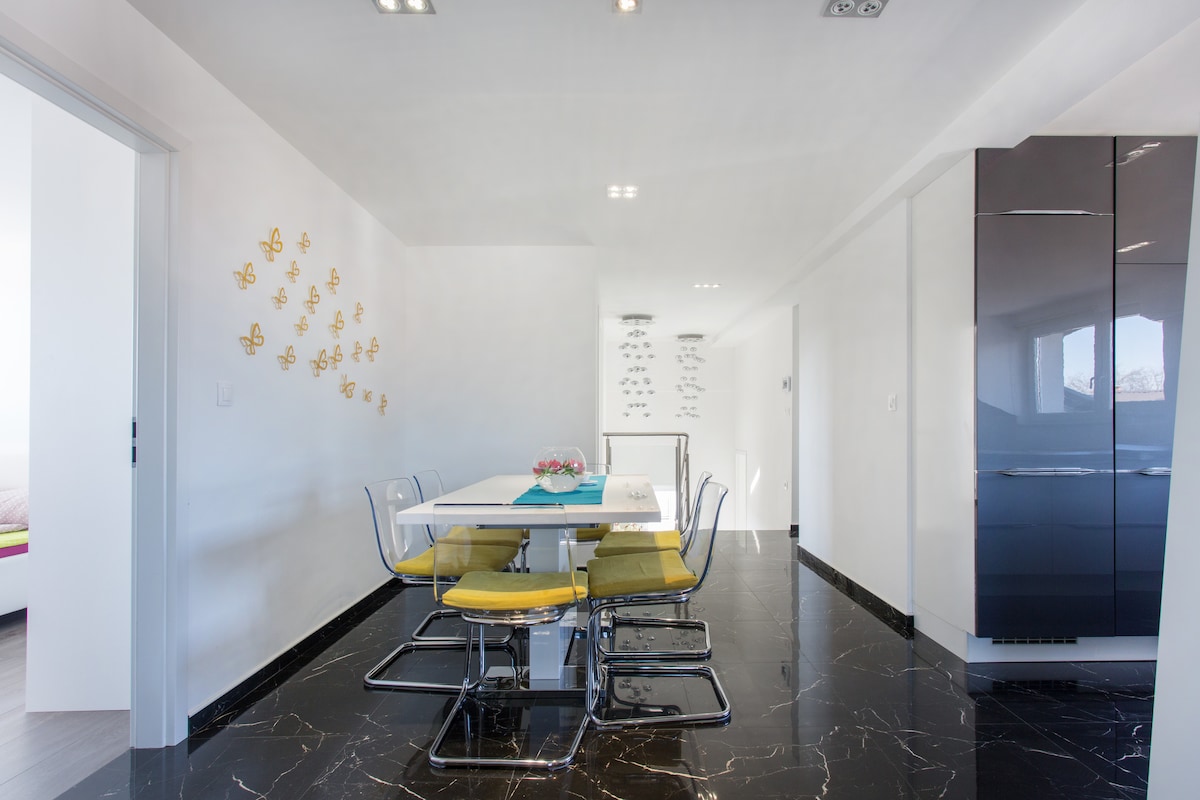 Dining area on black marble with the white gloss table and transparent chairs on the left, gold butterflies on the wall, and a tall dark gloss fridge-freezer standing against the right-hand wall