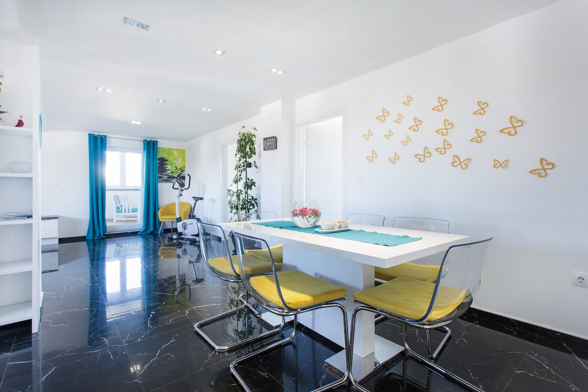 Wide view of the top-floor apartment: white walls and recessed downlights over a mirror-polished black marble floor, a white gloss dining table with yellow-cushioned transparent chairs, gold butterflies on the wall and turquoise curtains at the terrace door