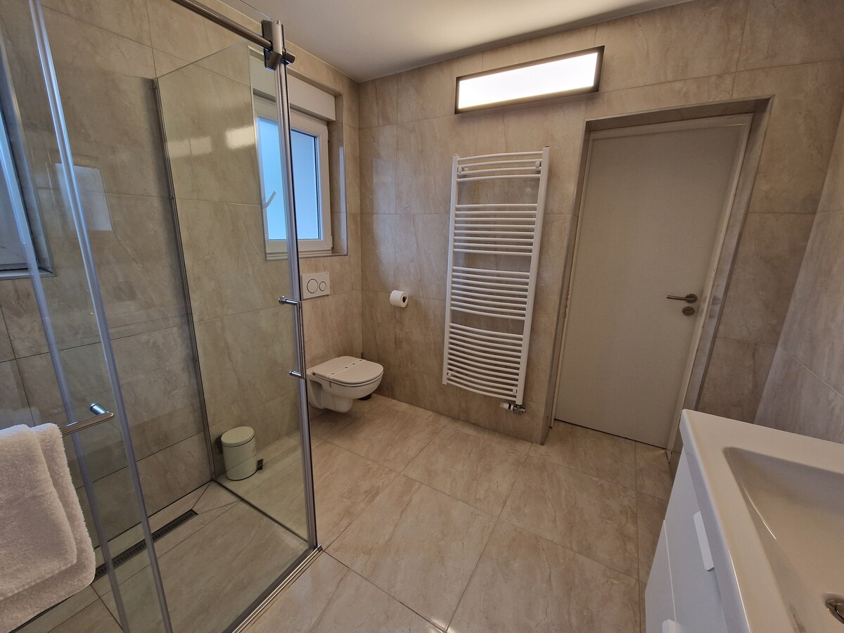 Doccia walk-in con parete in vetro accanto a un WC sospeso, con un termoarredo bianco alla parete e la porta del corridoio sulla destra