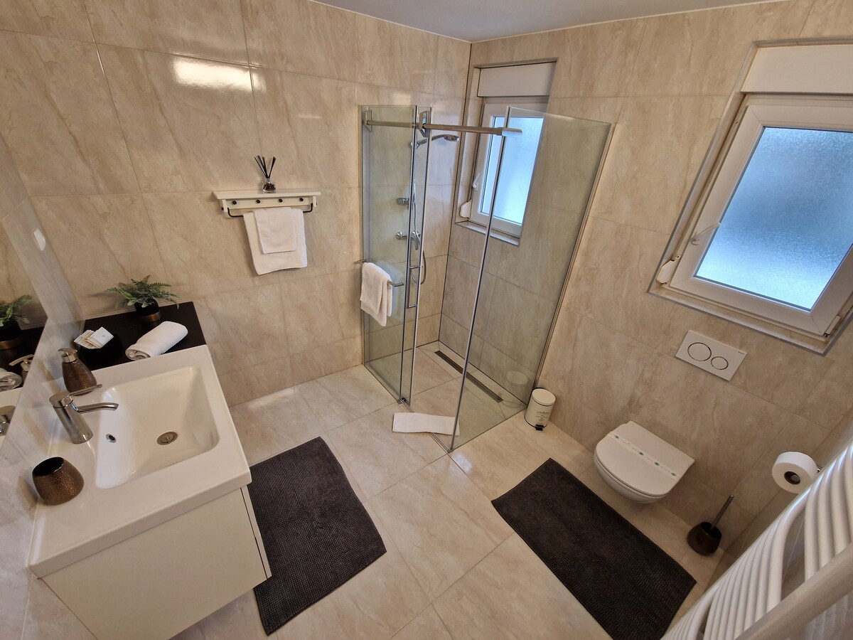 Bagno in piastrelle di pietra color crema con un lavabo bianco a sinistra, una doccia walk-in in vetro al centro, WC e bidet sospesi e tappetini scuri sul pavimento
