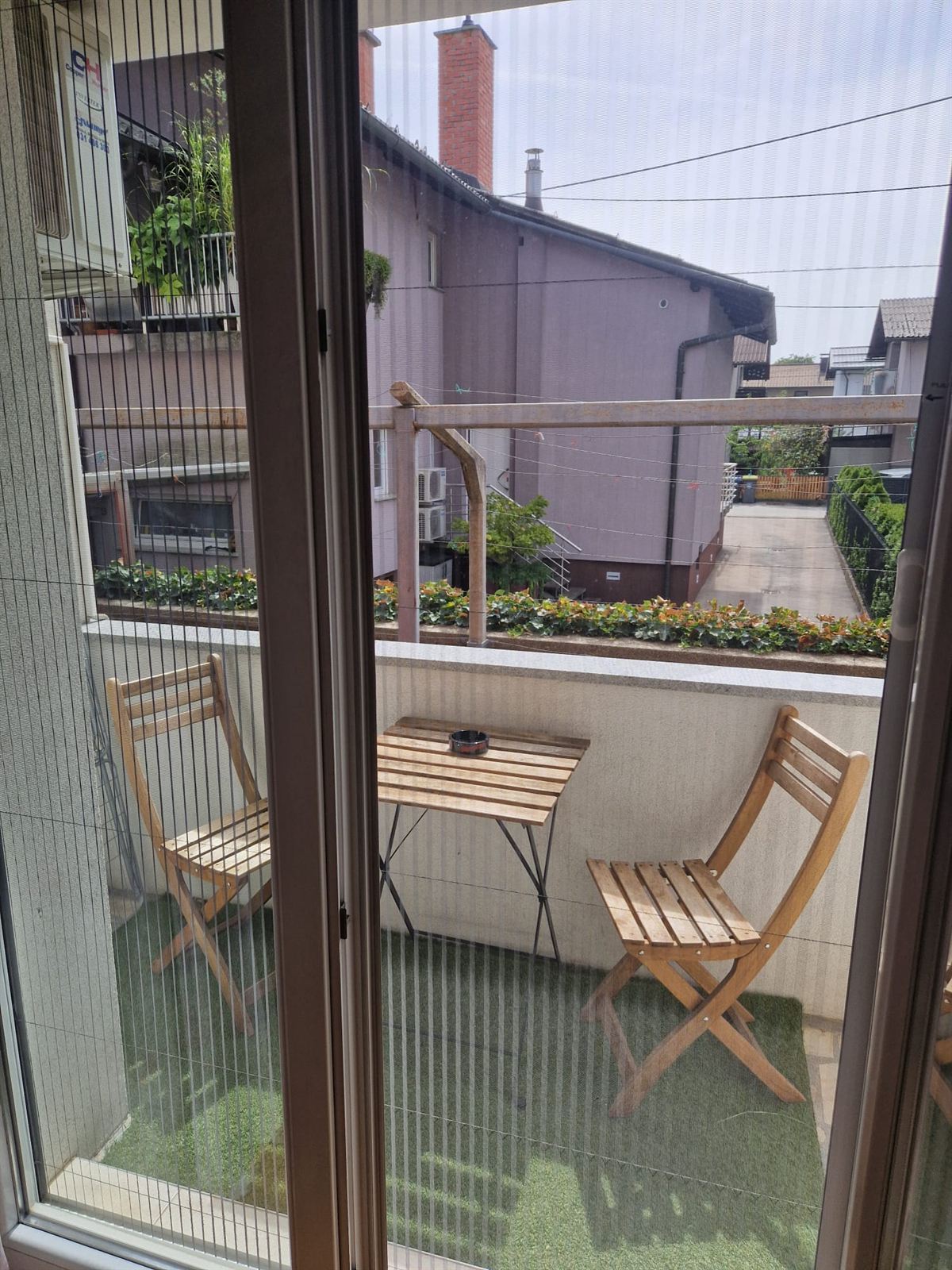 Vista attraverso la porta del balcone su un tavolino in legno e due sedie pieghevoli, con fioriere lungo la ringhiera