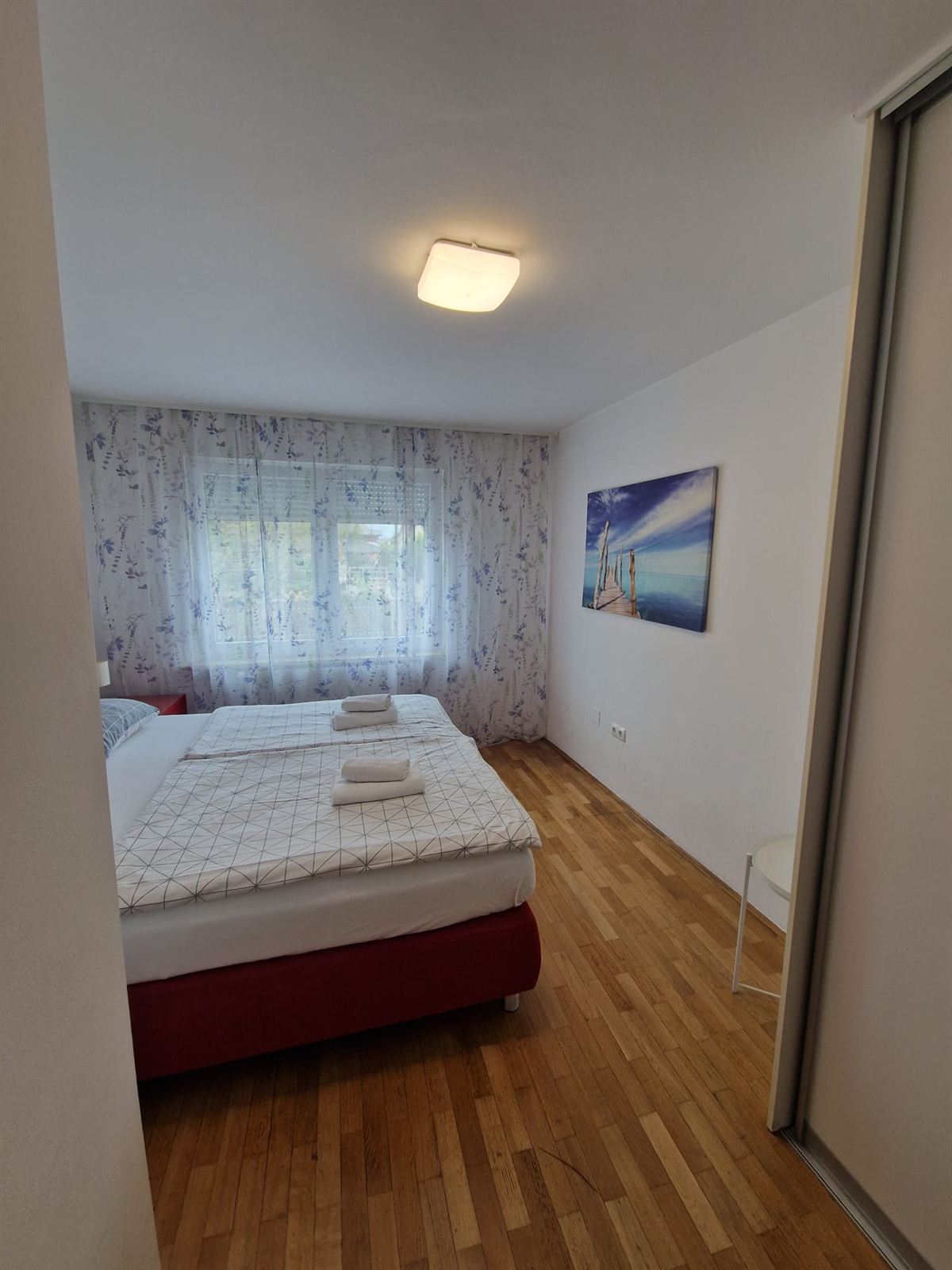 Camera con un letto matrimoniale su base rossa, una finestra con la tenda che lascia entrare la luce del giorno e un armadio scorrevole a specchio lungo la parete destra