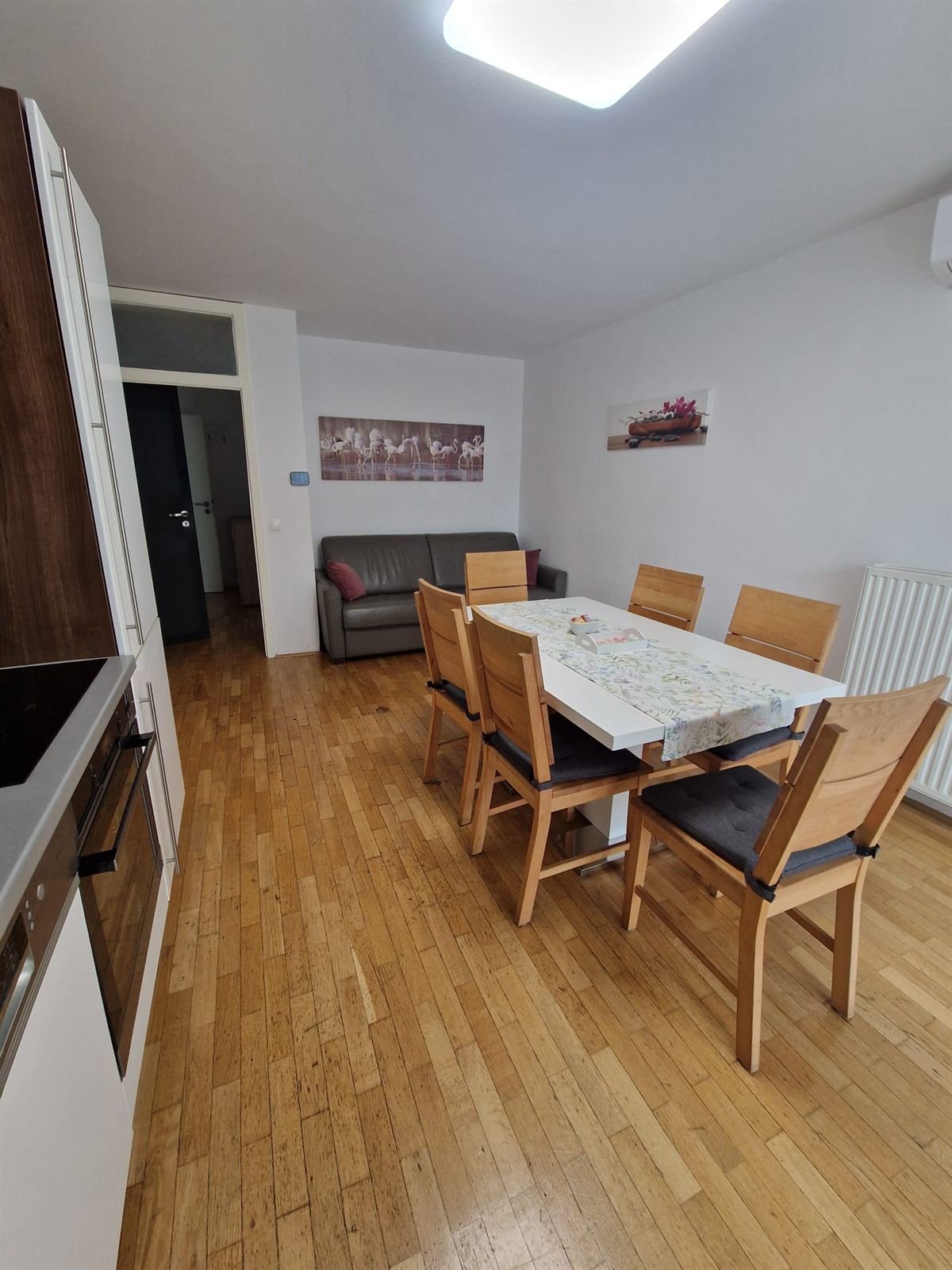 Un tavolo da pranzo con una tovaglia a fantasia e sei sedie in legno, un divano grigio e un radiatore in fondo alla stanza