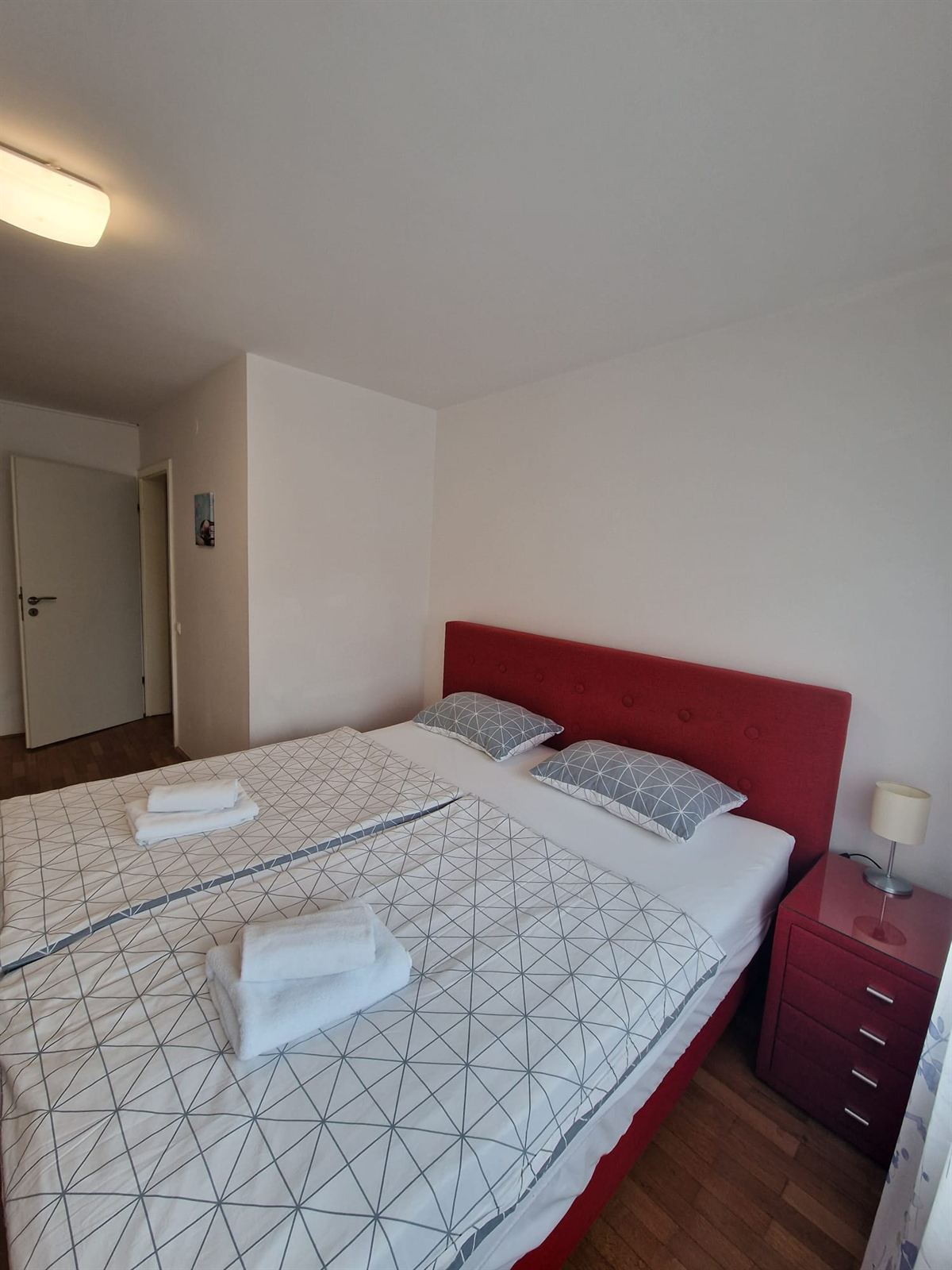 Letto matrimoniale con testiera imbottita rossa e biancheria grigia e bianca a motivo geometrico, un piccolo comodino rosso con una lampada, accanto a una porta bianca