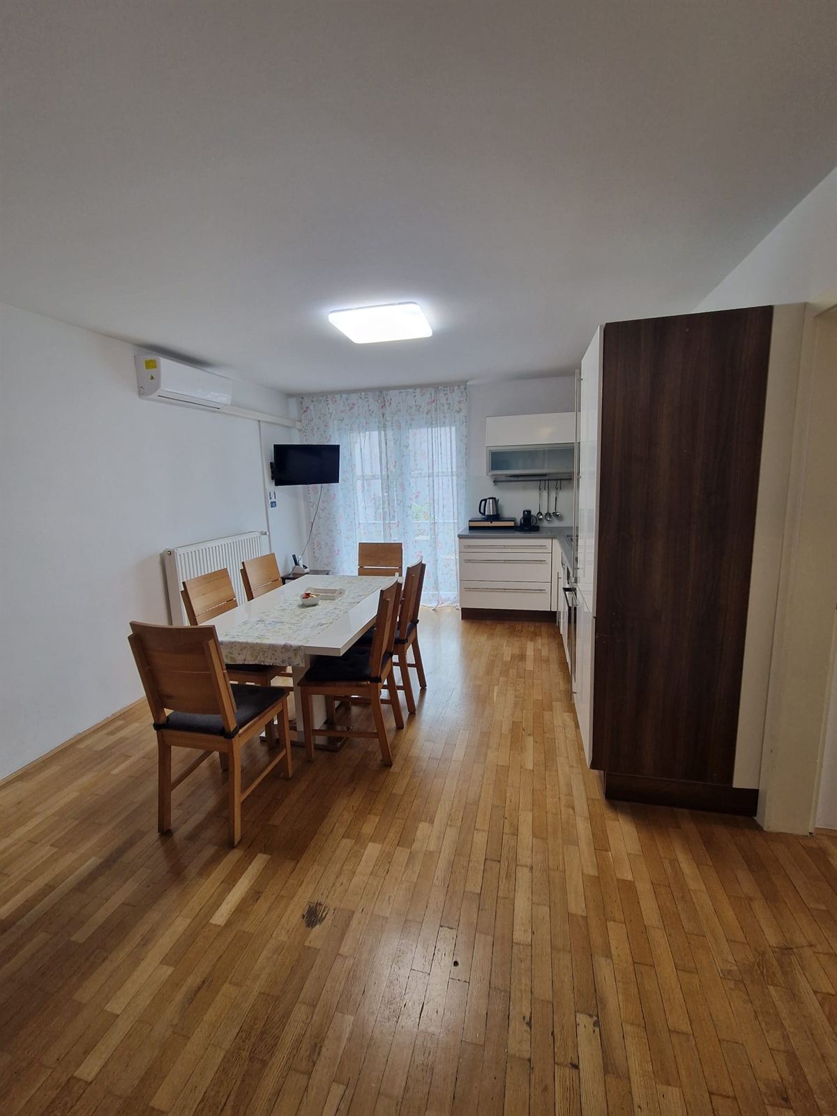 Glavna prostorija apartmana br. 1: duga otvorena prostorija s blagovaonskim stolom i šest stolica, kuhinjom na kraju i televizorom na zidu