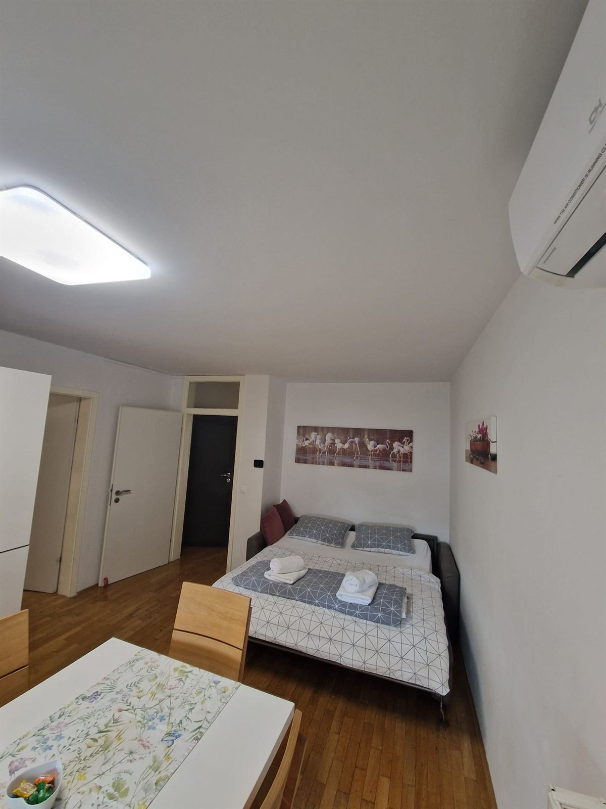 Ambiente open space sotto il tetto spiovente, con un divano letto già preparato sotto un'ampia tela con fenicotteri, un tavolo da pranzo in primo piano e una porta bianca verso il corridoio
