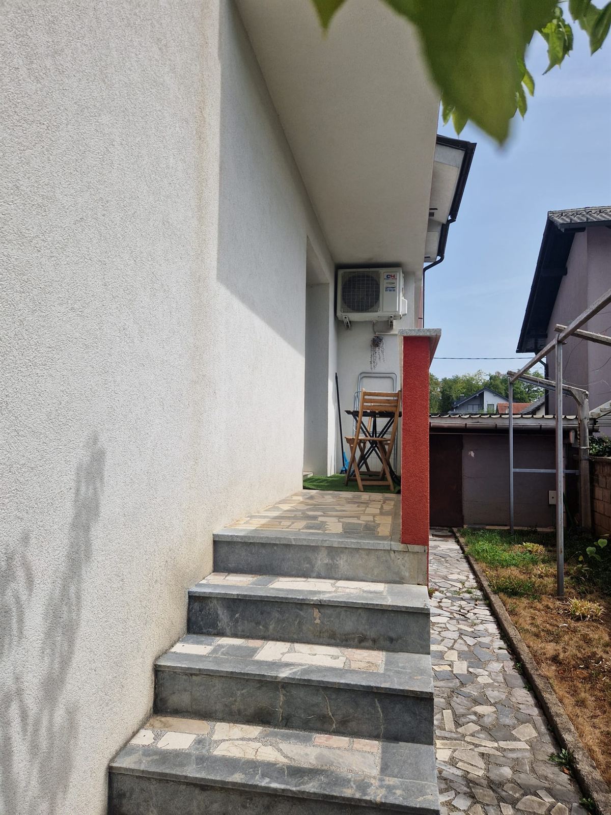 Gradini in pietra e piastrelle che salgono lungo il fianco della casa fino a un terrazzo con sedie pieghevoli, con un giardino e gli edifici vicini sullo sfondo