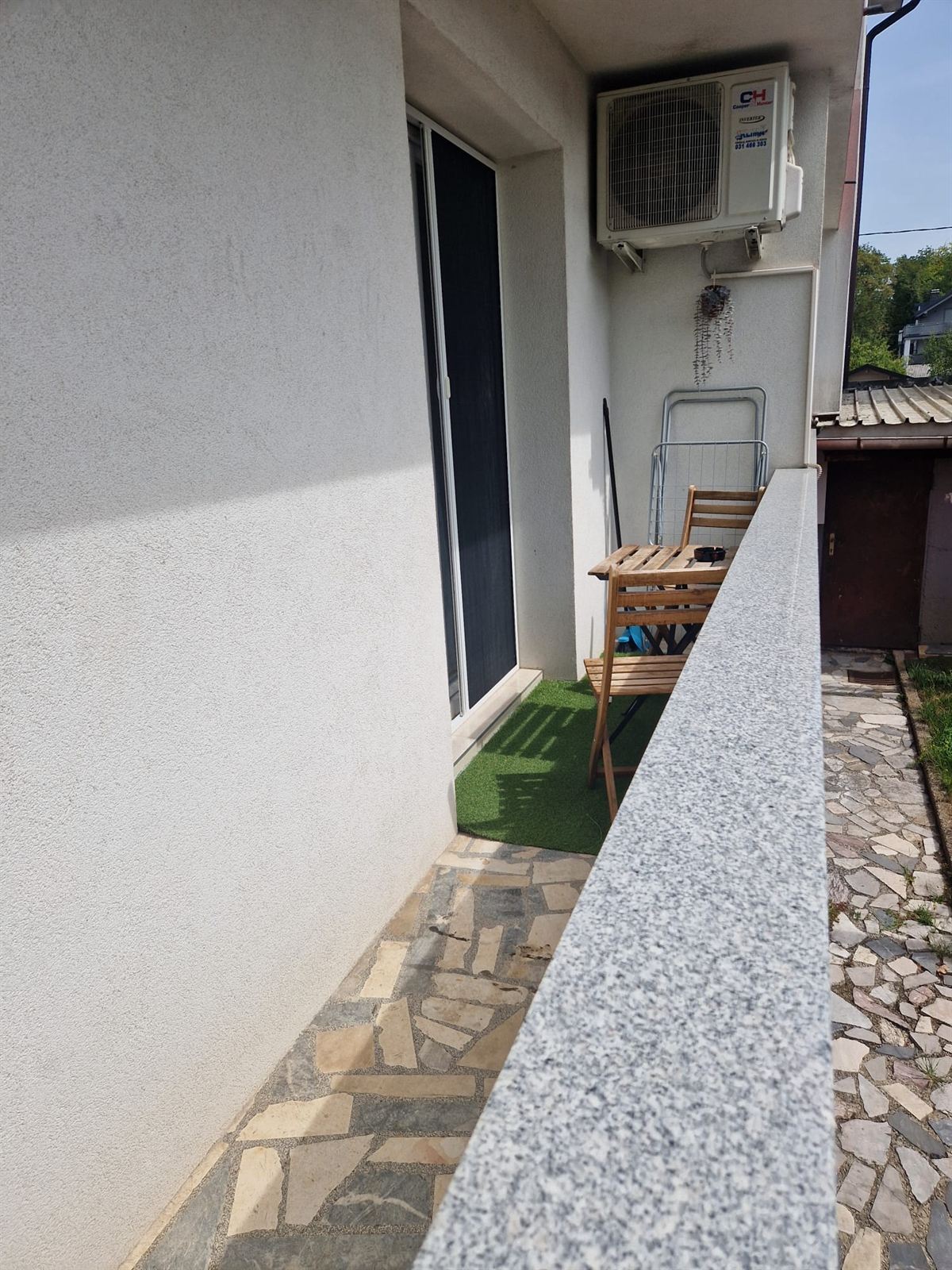 Un vialetto in pietra lungo la parete intonacata della casa fino a un piccolo terrazzo con tappeto verde e sedie pieghevoli in legno, con un condizionatore sulla parete sopra