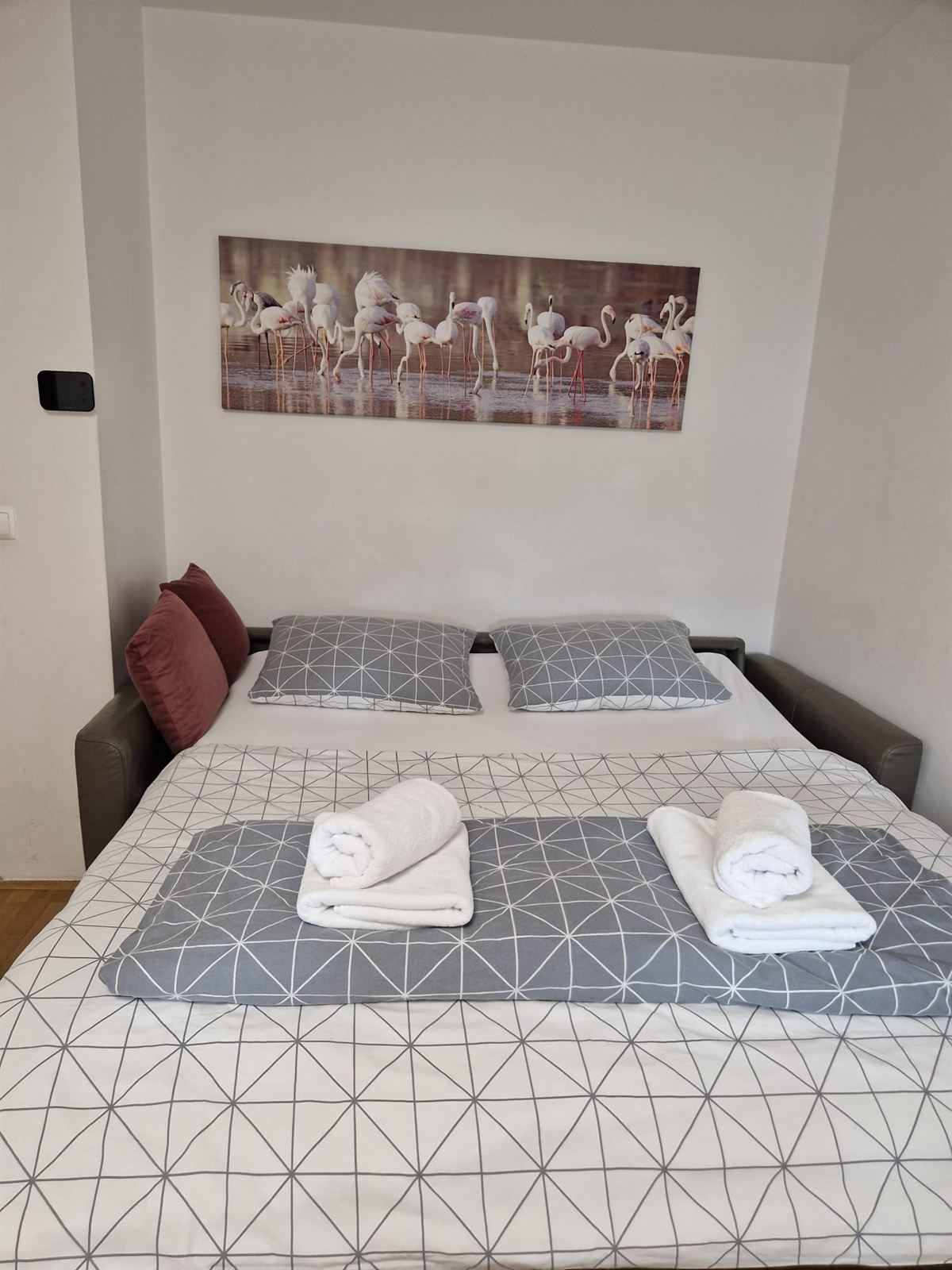 Un divano letto aperto e preparato con biancheria grigia e bianca a motivo geometrico, cuscini bordeaux sulla testata e asciugamani piegati sopra