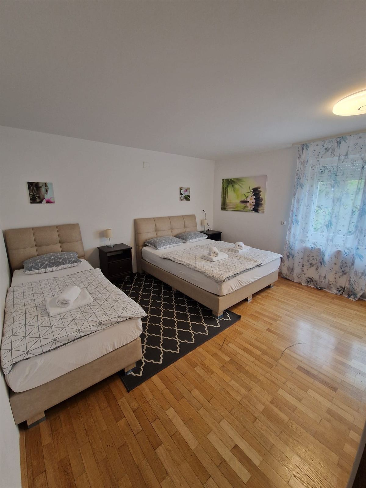 Camera con un letto matrimoniale e un letto singolo affiancati, testiere imbottite color crema, un tappeto a fantasia tra i due e una finestra con la tenda