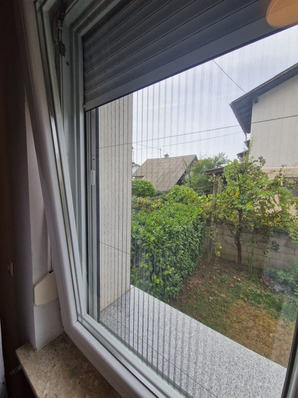 Vista da una finestra aperta con zanzariera su un giardino con alberi da frutto, un muretto in pietra e i tetti delle case vicine
